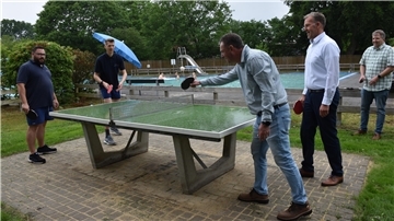 Fünf Männer spielen an einer Tischtennisplatte im Freien, im Hintergrund schwimmen Menschen in einem Freibad.