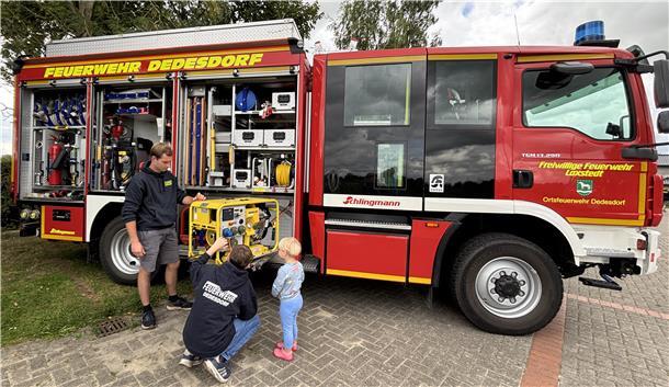 Zwei Feuerwehrleute der Ortsfeuerwehr Dedesdorf zeigen einem kleinen Kind technische Geräte am geöffneten Einsatzfahrzeug. Der Wagen ist mit Ausrüstung beladen und gut sichtbar beschriftet.