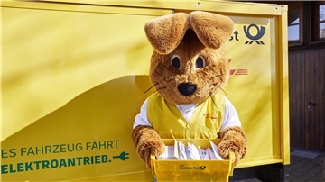 Ein Mensch in einem Hasenkostüm steht vor einem Postauto und hält eine Postkiste mit Briefen in den Pfoten.