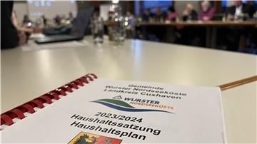 Im Vordergrund liegt der Haushaltsplan Hefter, im Hintergrund sind verschwommen Menschen an Tischen zu erkennen.