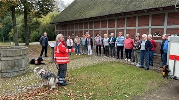 Eine Frau in roter Warnweste steht mit ihrem Hund vor einer Gruppe Menschen und erzählt etwas. 