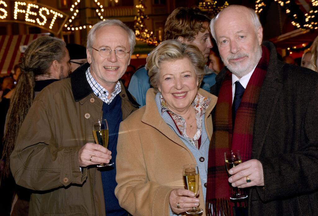 Die Schauspieler Joachim Hermann Luger (Hans Beimer, links) und die Schauspieler Marie-Luise Marjan (Mutter Beimer) und Bill Mockridge (Erich Schiller, rechts) posieren in der Außenkulisse mit Sektgläsern nach der Live-Ausstrahlung der 1559. Folge im Dezember 2015.