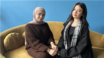 Zwei junge Frauen sitzen vor einer blauen Wand auf einem senfgelben Sofa. Die Frau links trägt ein braunes, langärmeliges Kleid und ein taupefarbenes Kopftuch. Die Frau rechts hat schwarze Haare, trägt einen schwarzen Mantel und einen karierten Schal.