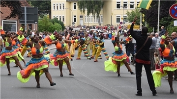 Tanzgruppe in bunten Kostümen zieht beim internationalen Trachtenfest durch eine Straße, begleitet von Musik und jubelnden Zuschauern am Straßenrand.