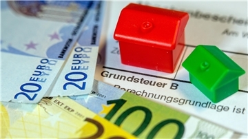 Mehrere Geldscheine, ein Grundsteuerbescheid und zwei kleine Holzhäuschen sind zu sehen.