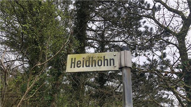Ein Straßenschild mit der Beschriftung "Heidhöhn" vor einigen Bäumen