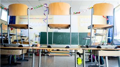 Mehrere Stühle stehen in einer leeren Schulklasse auf den Tischen. Im Hintergrund ist die Tafel zu erkennen.