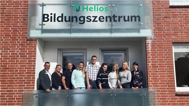 Eine Gruppe von neun Personen steht lächelnd auf dem Balkon des Helios Bildungszentrums vor einem Schild mit dem Schriftzug „Helios Bildungszentrum“.
