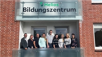 Eine Gruppe von neun Personen steht lächelnd auf dem Balkon des Helios Bildungszentrums vor einem Schild mit dem Schriftzug „Helios Bildungszentrum“.
