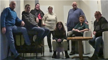Eine Gruppe von acht Personen, bestehend aus Männern und Frauen, sitzt und steht in einem Raum mit heller Wand und zwei geschlossenen Türen im Hintergrund. Einige Personen lehnen sich entspannt an Möbel, andere sitzen auf Stühlen oder hocken auf dem Boden.
