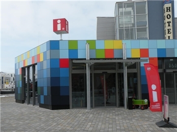 Ein buntes Gebäude in den Farben Grün, Gelb, Blau, Orange und Rot. Darüber ein roter Würfel, auf dem ein weißes "i" drauf steht. Er zeigt an, dass es sich um ein Info-Gebäude handelt. Das weiße "i" ist auch auf einer roten Fahne vor dem Gebäude zu sehen. 