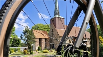 Ein Kirchengebäude mit rotem Ziegeldach und hohem Turm ist durch die Speichen eines Fahrrad-Vorderrads fotografiert. Im Vordergrund liegt ein gepflegter Friedhof.