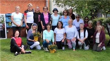 Eine Gruppe von Frauen unterschiedlichen Alters posiert fröhlich im Garten für ein Gruppenfoto. Einige knien oder sitzen im Gras, zwei halten Blumenschalen in den Händen. Im Hintergrund sind ein Apfelbaum, eine Hauswand und ein Spiegel zu sehen.