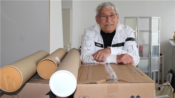 Künstler Hermy Eskorbier-Modersitzki lehnt sich in einem hellen Raum lächelnd auf einen Umzugskarton, daneben liegen mehrere Versandrollen mit Kunstwerken.