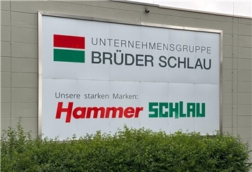 Das Logo der Unternehmensgruppe Brüder Schlau GmbH & Co KG und das des Raumaustatters Hammer ist auf einem Schild zu sehen.