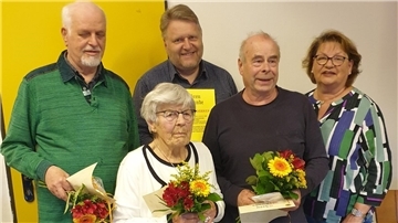 Fünf Personen, drei Männer und zwei Frauen, stehen zusammen. Vier von ihnen halten Blumensträuße und Urkunden in den Händen.