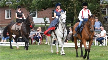 Drei Frauen in verschiedenen nostalgischen Outfits reiten auf ihren Pferden bei einer Vorführung auf dem Midlumer Markt. Im Hintergrund verfolgen Zuschauer das Geschehen vom Rand der Reitbahn.