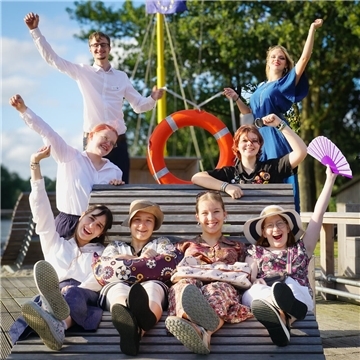 Sieben jungen Frauen und ein junger Mann sitzen und stehen rund um eine Sitzbank am Wasser. Sie lächeln und jubeln in die Kamera.