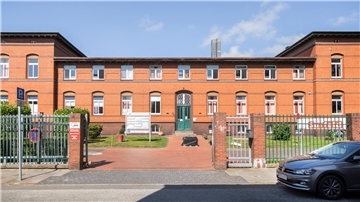 rotes Backsteingebäude mit grüner Tür in einer parkähnlichen Anlage hinter einem Zaun