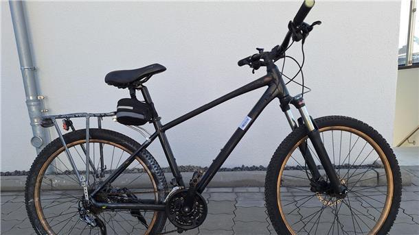 Ein schwarzes Fahrrad mit goldfarbenen Felgen steht vor einer weißen Wand. 