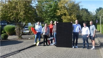 Sieben Männer stehen auf einer gepflasterten Straße und halten ein Photovoltaik-Modul in ihrer Mitte.