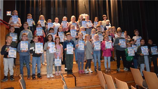 Dutzende Kinder stehen auf einer Holztribüne. In ihren Händen halten sie jeweils eine Urkunde. 