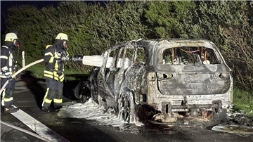 Zwei Feuerwehrleute stehen neben einem ausgebrannten Auto und besprühen es mit Löschschaum.