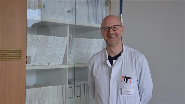 Ein Arzt steht in weißem Kittel vor einem Bücherregal und lächelt in die Kamera.