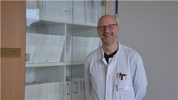 Ein Arzt steht in weißem Kittel vor einem Bücherregal und lächelt in die Kamera.