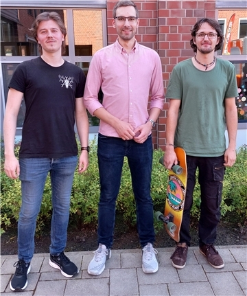 Drei junge Männer stehen nebeneinander vor einem Backsteingebäude und lächeln in die Kamera. Der Mann rechts hält ein Skateboard mit buntem Aufdruck in der Hand. Alle drei wirken entspannt und lässig gekleidet.