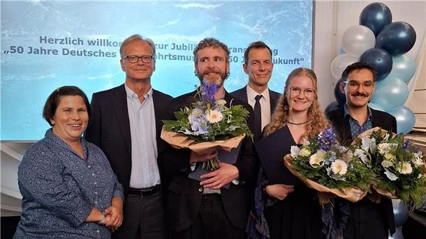 Gruppenfoto bei der Jubiläumsveranstaltung „50 Jahre Deutsches Schifffahrtsmuseum“. Drei ausgezeichnete Personen halten Blumensträuße, umgeben von Gratulanten. Im Hintergrund Ballondekoration und eine Leinwand mit Veranstaltungsaufschrift.
