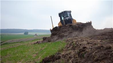 Ein Bagger schält den Mutterboden vom Deich ab. Anschließend wird er mit schwerem Kleiboden erhöht. Am Ende wird der Mutterboden wieder breitgefahren, damit das Gras schneller anwächst.