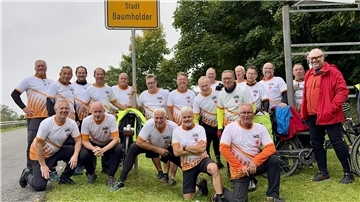 Eine Gruppe von 19 Radfahrern in einheitlichen Trikots steht lächelnd unter dem Ortsschild der Stadt Baumholder. Im Hintergrund sind Bäume und mehrere Fahrräder zu sehen.