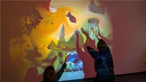 Wochenende voller Musik und Fantasie – „Mukkekukke“ lädt Kinder ein Eine Frau und ein Kind stehen vor einem magisch leuchtenden Wandbild und werfen Schatten darauf.