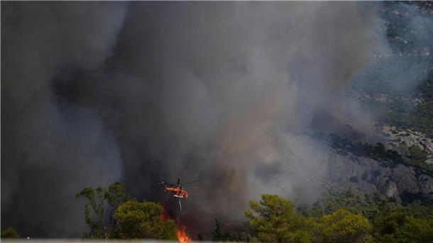 Brände in Griechenland „größte in der Geschichte der EU“ Ein Feuerwehrhubschrauber wirft Wasser auf die Flammen nahe des Militärstützpunkts Kapota im Gebirge Parnitha.