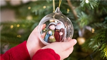 Ein Kind hält eine Krippe mit Maria, Josef und dem Jesuskind in einer gläsernen Weihnachtskugel an einem Weihnachtsbaum.
