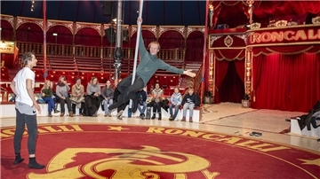 Ein Mann sitzt auf einer Pole-Stange, die durch die Luft schwingt.  Andere Personen schauen ihn an.