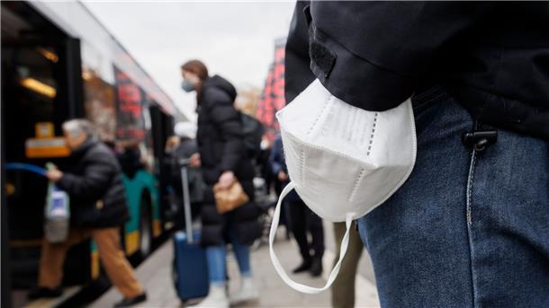 ÖPNV-Maskenpflicht fällt in zwei Bundesländern  Ein Mann wartet mit einer Maske am Handgelenk auf einen Bus.