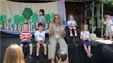 Eine Gruppe von Kindern und eine Erzieherin sitzen auf einer Bühne vor einem gemalten Waldhintergrund. Ein Mädchen im Rollstuhl ist ebenfalls Teil der Gruppe. Alle Beteiligten wirken fröhlich und entspannt bei sommerlichem Wetter.