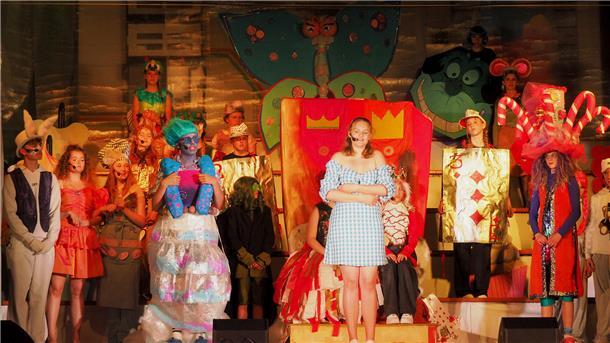 Schülerinnen und Schüler des Gymnasiums Wesermünde in bunten Kostümen auf der Bühne während des Musicals „Alice“. Im Mittelpunkt steht eine Schülerin im blau-weißen Kleid als Alice, umgeben von fantasievoll verkleideten Figuren wie Karten, Tieren und Fabelwesen vor einer farbenfrohen Kulisse.
