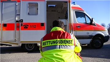 Ein Mann trägt eine Jacke mit der Aufschrift  ,,Rettungsdienst". Dahinter steht ein Krankenwagen mit geöffneter Tür.
