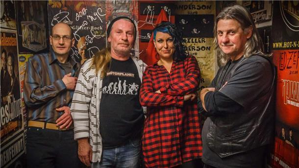 Bandfoto von Motorplanet: Drei Männer und eine Frau stehen in einem Raum mit vielen Bandplakaten.