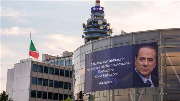 Eine italienische Flagge auf Halbmast weht vor einer Nachricht mit der Aufschrift „Grazie Silvio“ auf einem Turm des Fernsehsenders Mediaset neben einem großen Plakat.