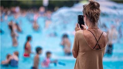 Eine junge Frau fotografiert mit ihrem Handy in einem Freibad. Hunderttausende Kinder und Erwachsene folgen minderjährigen Influencern auf Youtube, Instagram oder TikTok durch ihren Alltag, in den Park oder ins Schwimmbad. Hinter der Kamera, den Accounts und den Verträgen mit Unternehmen, die in den Videos ihre Produkte platzieren, stehen oft die Eltern.