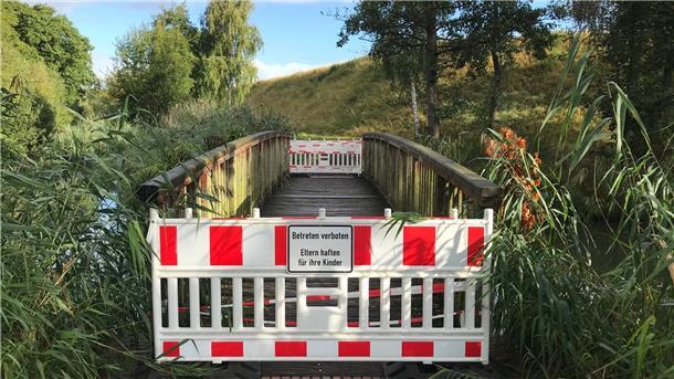 Über diese Brücke kann man derzeit nicht mehr gehen Eine kleine Fußgängerbrücke führt, tritt man durch das einstige Tor der Admiral-Brommy-Kaserne, in eine Grünanlage mit Wegen. Die Brücke ist derzeit gesperrt. Ihr Belag muss ersetzt werden.