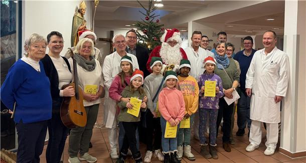 Eine fröhliche Gruppe aus Erwachsenen und Kindern steht vor einem festlich geschmückten Weihnachtsbaum in einem Krankenhaus. Einige Erwachsene tragen Arztkittel, andere sind festlich gekleidet, darunter auch ein Weihnachtsmann mit rotem Mantel und Bart. Die Kinder tragen Weihnachtsmützen oder grüne Zipfelmützen und halten gelbe Hefte in den Händen.