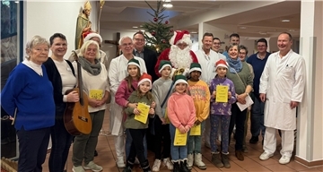 Eine fröhliche Gruppe aus Erwachsenen und Kindern steht vor einem festlich geschmückten Weihnachtsbaum in einem Krankenhaus. Einige Erwachsene tragen Arztkittel, andere sind festlich gekleidet, darunter auch ein Weihnachtsmann mit rotem Mantel und Bart. Die Kinder tragen Weihnachtsmützen oder grüne Zipfelmützen und halten gelbe Hefte in den Händen.