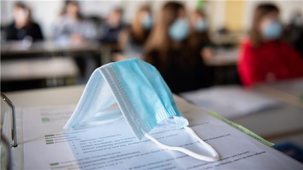 Impfung von Kindern ab 12 vor neuem Schuljahr angepeilt Eine Maske liegt im Unterricht auf Unterlagen, während im Hintergrund Schülerinnen und Schüler in einem Gymnasium mit Mund- und Nasenschutz zu sehen sind. Für Kinder ab 12 könnten Impfungen dieses Bild verhindern.