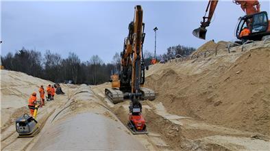 Eine Rüttelplatte wird von einem Bauarbeiter gezogen, die andere von einem Bagger: Schicht für Schicht wird der Sand um das neue Rohr verdichtet.
