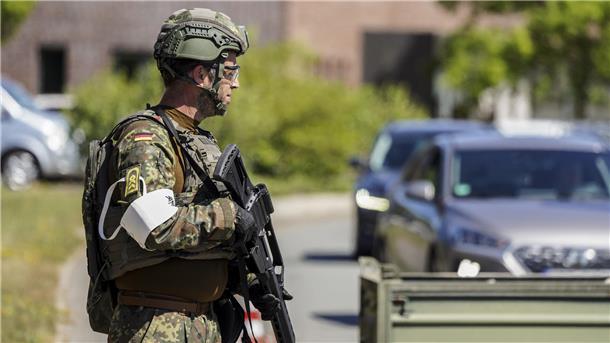Ein Bundeswehrsoldat in Tarnuniform und Schutzhelm steht mit Gewehr an einer Straße. Im Hintergrund warten Autos vor einer Kontrollstelle.